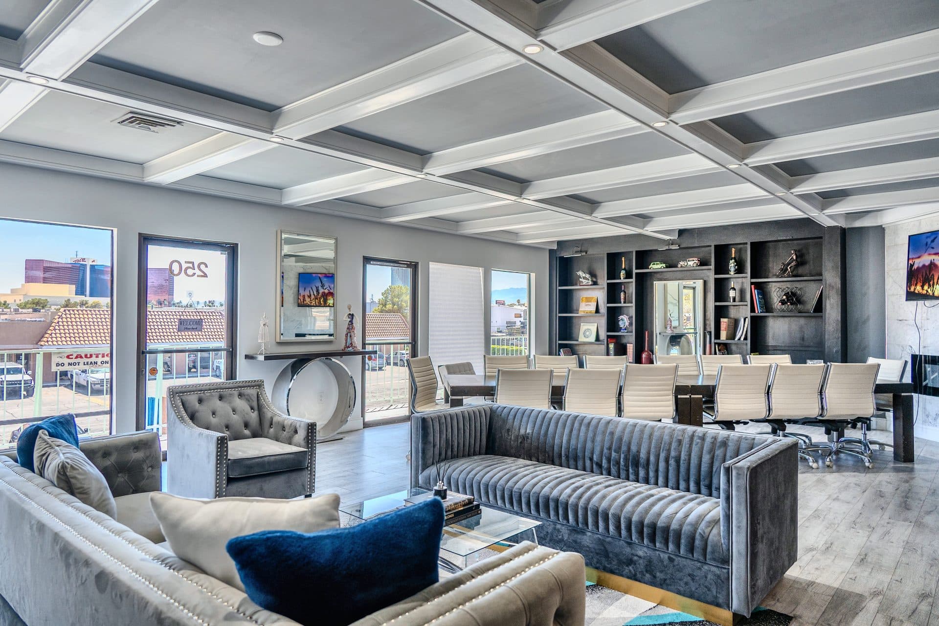 The Library Lounge: gray velvet sofas with blue cushions under a coffered ceiling, a long table with cream chairs, dark bookshelves and a marble fireplace wall
