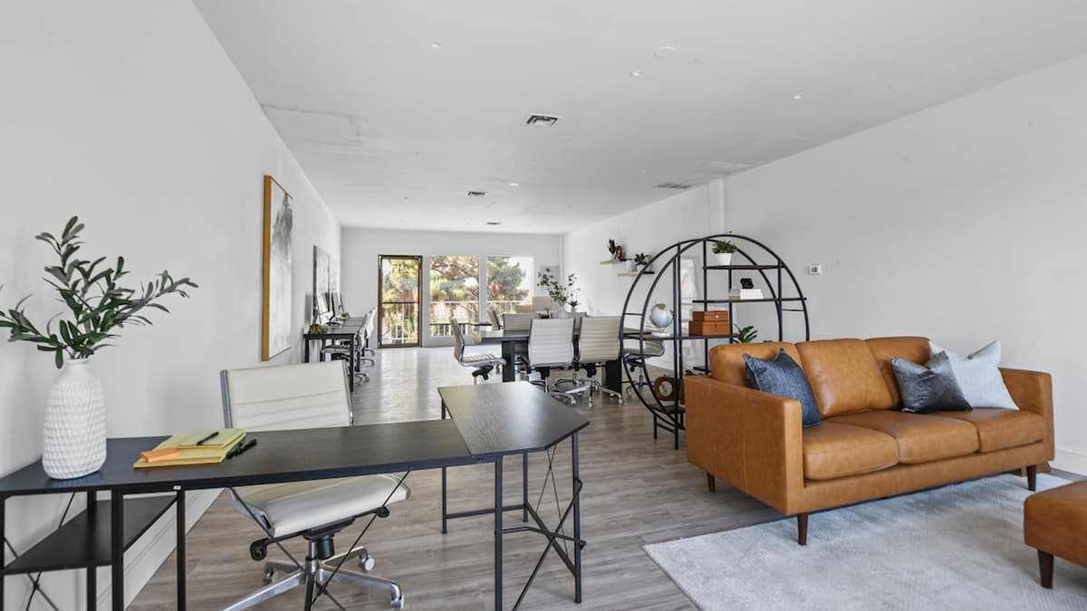 Espacio de trabajo abierto con un escritorio negro, una silla de cuero blanco, un sofá de cuero color coñac y una mesa compartida junto a las ventanas