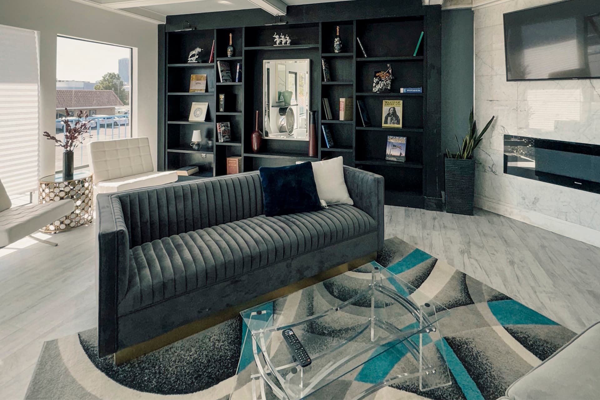 Library-style lounge with a gray velvet sofa, black built-in bookshelves, a marble fireplace wall and a TV